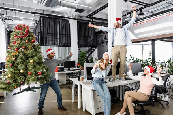 Alegre empresario indio sosteniendo árbol de Navidad cerca de colegas con copas de champán en la oficina - foto de stock