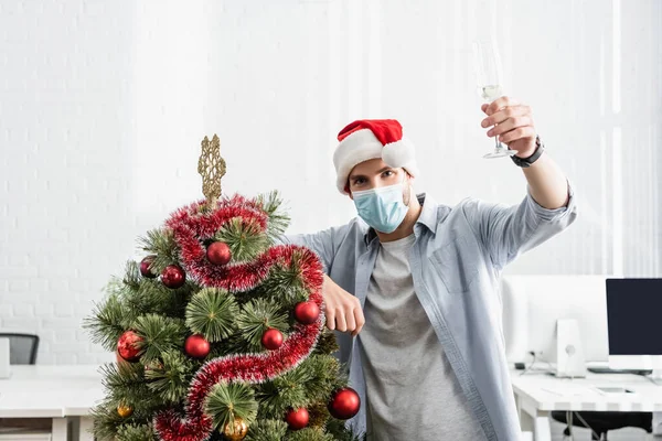 Бизнесмен в медицинской маске и шляпе Санты с бокалом шампанского возле рождественской елки в офисе — стоковое фото