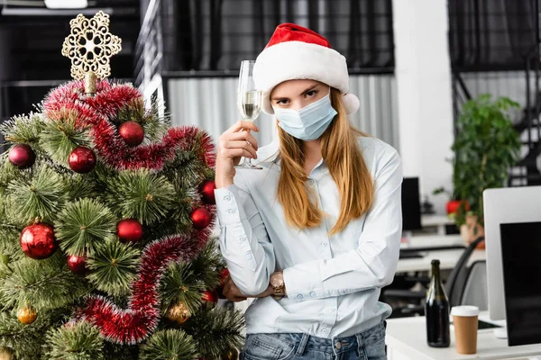Empresaria en máscara médica sosteniendo copa de champán cerca del árbol de Navidad en la oficina - foto de stock
