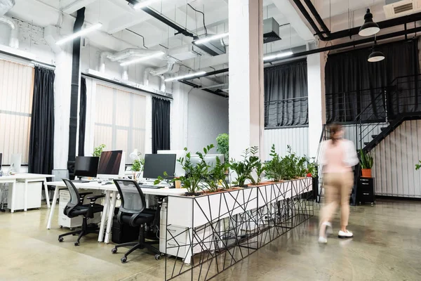 Desenfoque de movimiento de la mujer de negocios caminando cerca de computadoras y plantas en la oficina - foto de stock