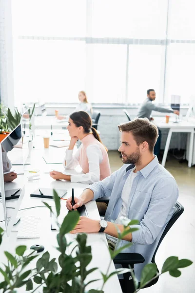 Businessman using graphics tablet and computer near colleagues on blurred background in office — Stock Photo