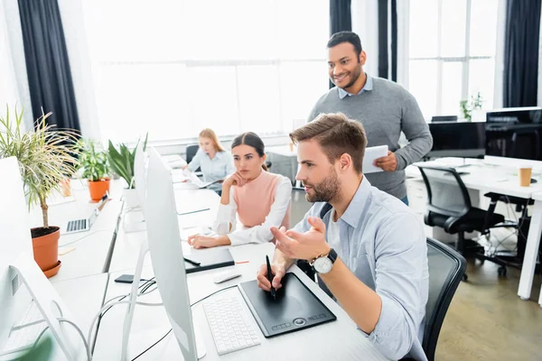 Businessman using graphics tablet and computer near multicultural colleagues in office — Stock Photo