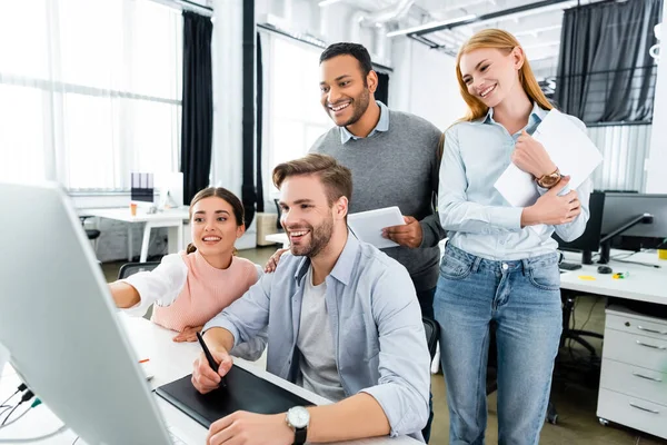 Alegre gente de negocios multiétnicos de pie cerca de colega utilizando la computadora y la tableta gráfica en la oficina - foto de stock