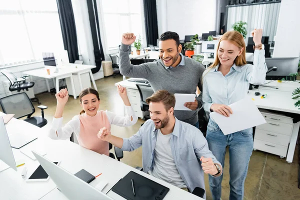 Negocios multiétnicos alegres con tableta digital y papeles de pie cerca de la computadora en la oficina - foto de stock