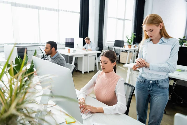 Businesswoman with papers looking at wristwatch near colleague using computer in office — Stock Photo