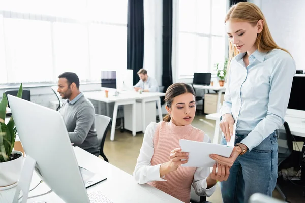 Geschäftsfrauen schauen sich Dokumente in der Nähe des Computers an und indische Kollegen auf verschwommenem Hintergrund im Büro — Stockfoto