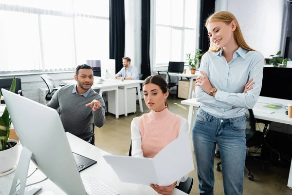 Empresaria mirando papeles cerca de colegas multiétnicos y computadora en la oficina - foto de stock