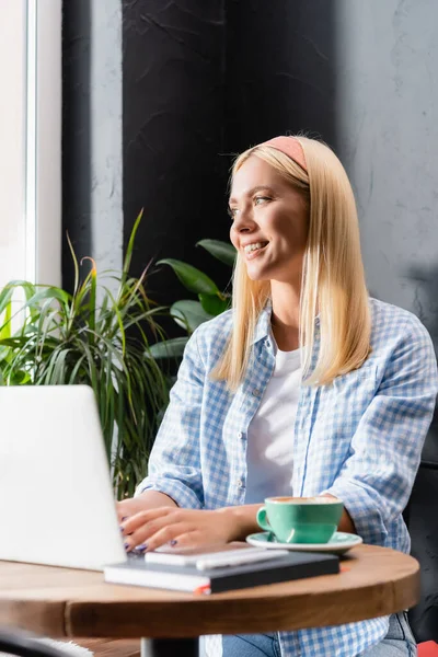Blonde freelancer smiling while typing on laptop in cafe on blurred foreground — Stock Photo