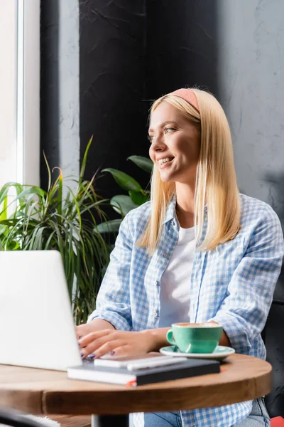 Blondine Freiberuflerin lächelt beim Tippen auf Laptop im Café im verschwommenen Vordergrund — Stockfoto