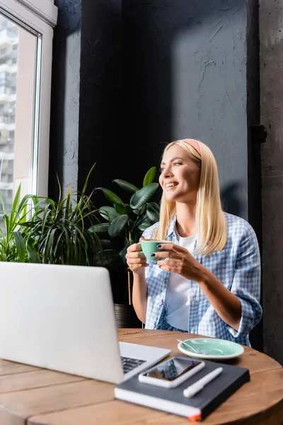 Freelancer feliz segurando xícara de café perto do laptop em primeiro plano borrado — Fotografia de Stock