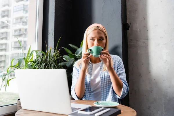 Rubia freelance beber café cerca de la computadora portátil en la cafetería, borrosa primer plano - foto de stock