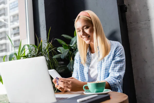 Alegre rubia freelancer en camisa a cuadros charlando en el teléfono inteligente cerca de la taza de café y portátil en primer plano borrosa - foto de stock