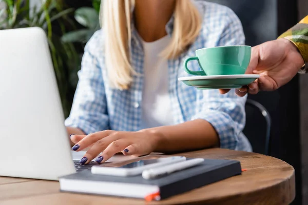 Cropped view of waiter holding cup of coffee near freelancer typing on laptop in cafe — Stock Photo
