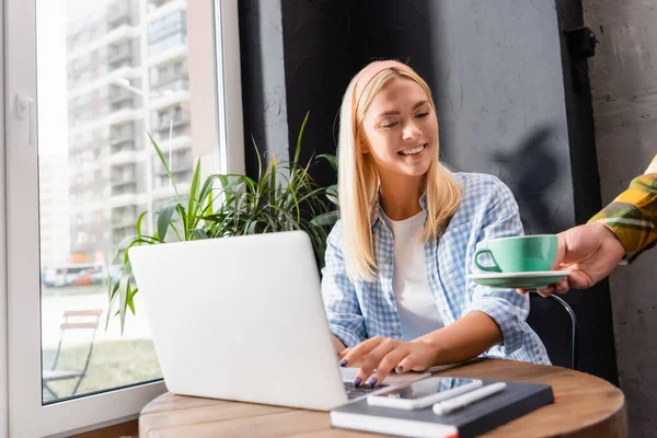Freelancer sorrindo digitando no laptop enquanto garçom servindo café, foreground borrado — Fotografia de Stock