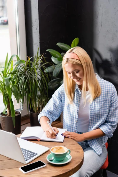 Scrittura freelance sorridente in notebook vicino al computer portatile, smartphone con schermo bianco e tazza di caffè — Foto stock