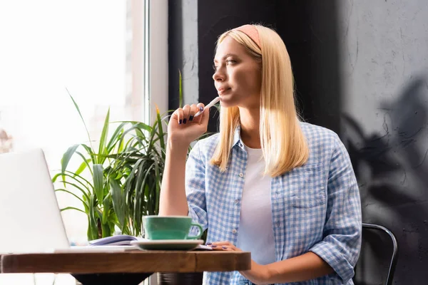 Nachdenkliche Freiberuflerin hält Stift in der Hand, während sie im Café neben Laptop und Tasse Kaffee sitzt — Stockfoto