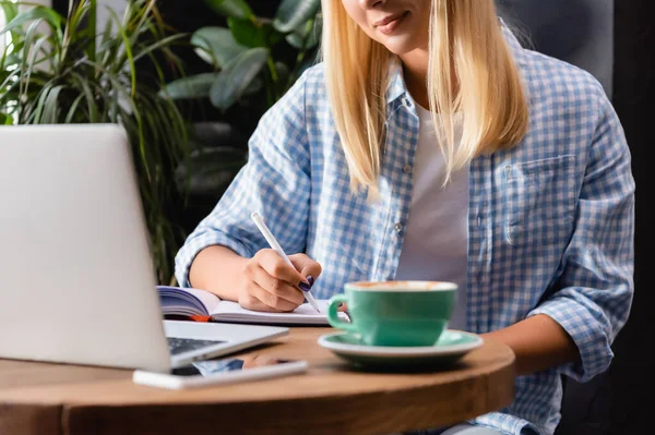 Ausgeschnittene Ansicht eines Freiberuflers im karierten Hemd, der in Notizbuch neben Tasse Kaffee und Laptop auf verschwommenem Vordergrund schreibt — Stockfoto