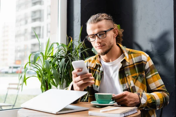 Стильний фрілансер у простій сорочці та окулярах спілкується на смартфоні біля чашки кави та ноутбука, розмитий передній план — стокове фото