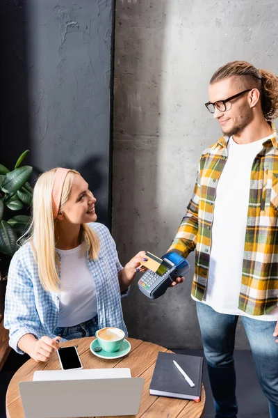 Lächelnder Kellner mit Zahlungsterminal in der Nähe der blonden Freiberuflerin, die in einem Café in der Nähe von Gadgets sitzt — Stockfoto