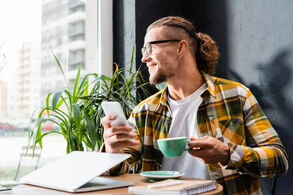 Lächelnder Freiberufler mit Brille und kariertem Hemd, Kaffeetasse und Smartphone in der Hand, während er neben Laptop im Café sitzt — Stockfoto