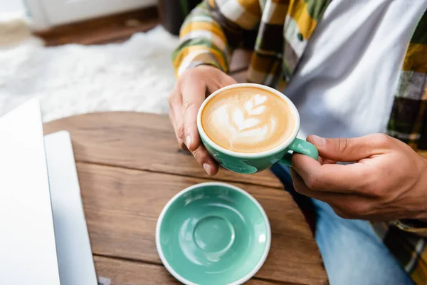 Cropped view of man sitting in cafe and holding cup of coffee with latte art — Stock Photo