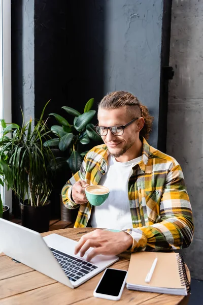 Stylish freelancer in checkered shirt and eyeglasses using laptop while holding cup of coffee in cafe — Stock Photo
