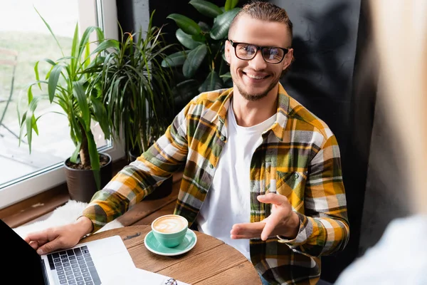 Joyeux pigiste dans les lunettes et chemise à carreaux pointant avec la main près de tasse de café et ordinateur portable dans le café — Photo de stock