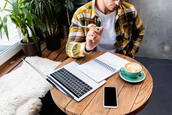 Vista ritagliata di freelance in caffè vicino a computer portatile, notebook, tazza di caffè e smartphone con schermo bianco — Foto stock
