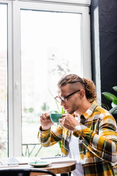 Pigiste élégant dans des lunettes et chemise à carreaux tenant tasse de café tout en étant assis dans un café près d'un ordinateur portable — Photo de stock