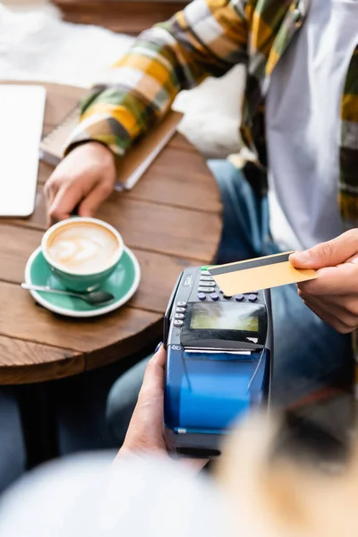 Обрезанный вид официанта, держащего платежный терминал, и человека с кредитной картой, сидящего за столом с чашкой кофе — стоковое фото