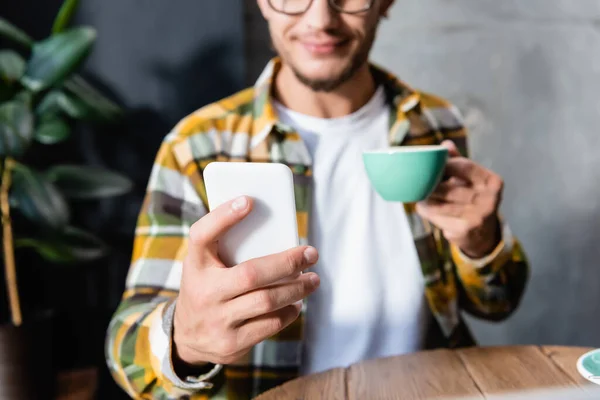 Partial view of freelancer messaging on smartphone while holding cup of coffee, blurred background — Stock Photo