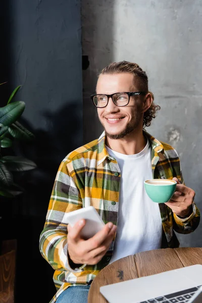 Felice freelance in camicia a quadri e occhiali distogliendo lo sguardo tenendo smartphone e tazza di caffè in primo piano sfocato — Foto stock