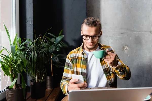 Elegante freelance in camicia a quadri e occhiali che chatta su smartphone e tiene la tazza di caffè vicino al computer portatile in primo piano sfocato — Foto stock