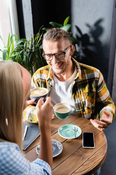 Sorridente giovane uomo che tiene in mano una tazza di caffè mentre parla con una donna nel caffè — Foto stock