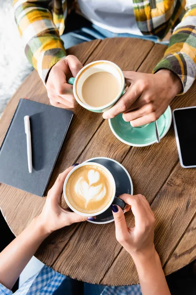 Top view of woman holding cup of coffee with latte art near friend in cafe — Stock Photo