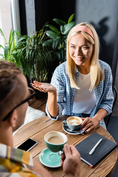 Joven mujer sonriente hablando con un amigo sosteniendo una taza de café en un primer plano borroso - foto de stock