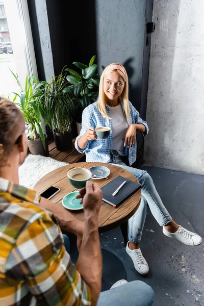 Smiling blonde woman holding cup of coffee while sitting with friend in cafe — Stock Photo