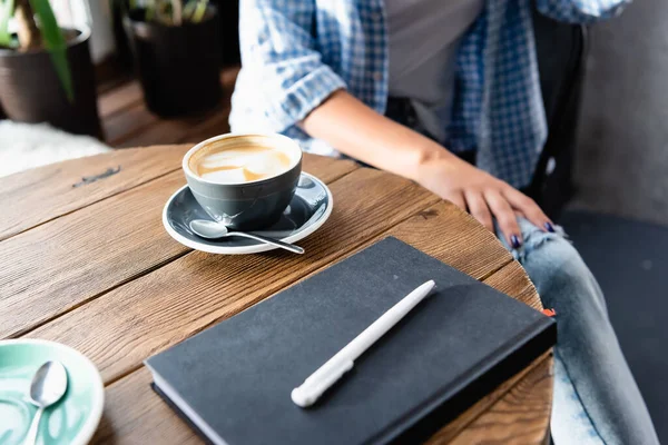 Vista ritagliata della donna seduta a tavola con tazza di caffè e taccuino nel caffè — Foto stock