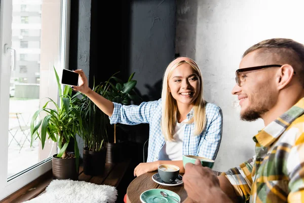 Счастливая блондинка делает селфи на смартфоне с улыбающимся мужчиной, держащим чашку кофе на размытом переднем плане — стоковое фото