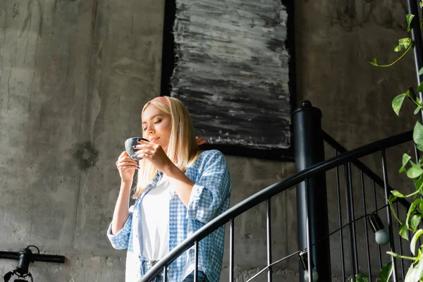 Young blonde woman in plaid shirt enjoying flavor of coffee while holding cup on stairway in cafe — Stock Photo