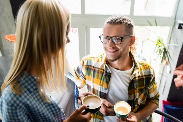 Giovane uomo sorridente in occhiali e donna in primo piano offuscata che tiene tazze di caffè in caffè — Foto stock