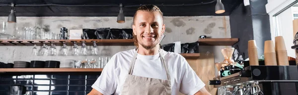 Smiling barista in apron looking at camera at workplace in coffee house, banner — Stock Photo