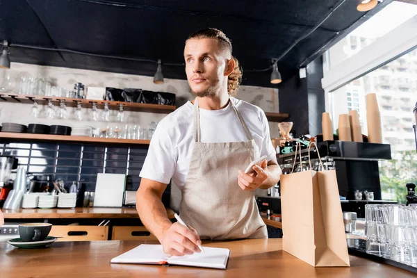 Junger Barista hält Smartphone beim Schreiben in Notizbuch neben Papiertüte auf Theke — Stockfoto