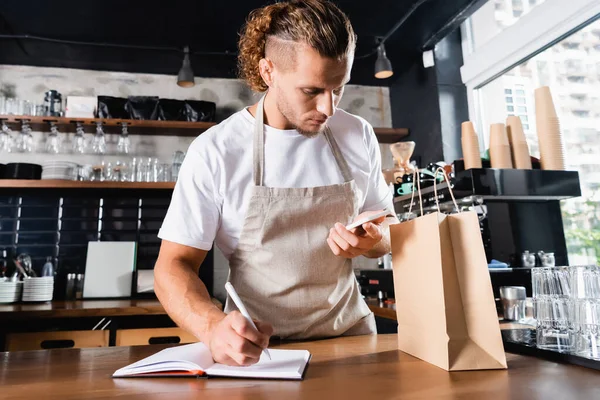 Bonito, jovem barista em avental escrevendo em notebook enquanto olha para smartphone perto de saco de papel no balcão bar — Fotografia de Stock