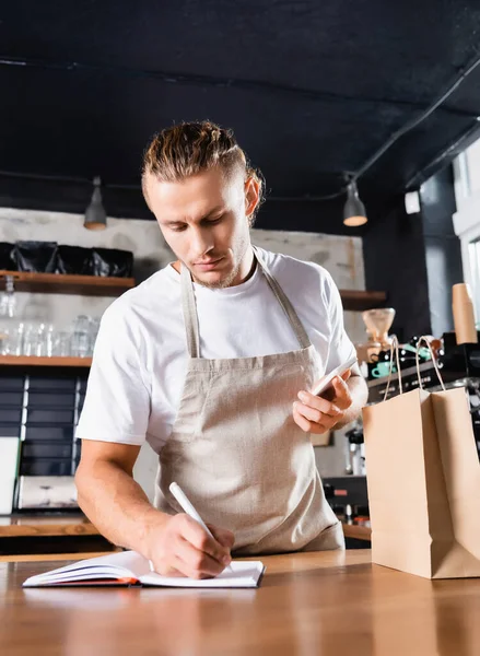 Young barista holding mobile phone and writing in notebook near paper bag on bar counter — Stock Photo