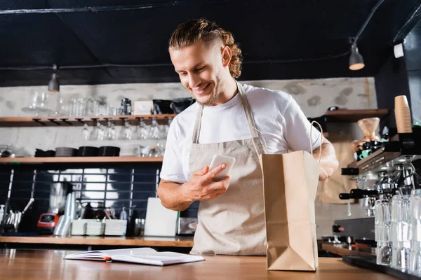 Barista sonriente en delantal mirando en el cuaderno mientras sostiene el teléfono inteligente cerca de la bolsa de papel en el mostrador de la barra - foto de stock