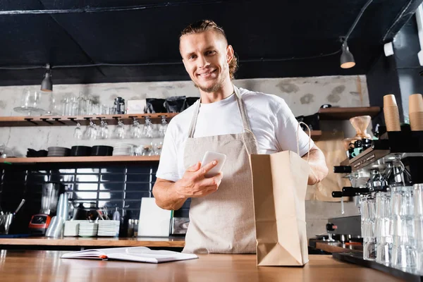 Smiling barista in apron holding mobile phone near notebook and paper bag on bar counter — Stock Photo