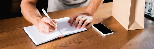 Partial view of waiter writing in notebook near mobile phone with blank screen on bar counter, banner — Stock Photo