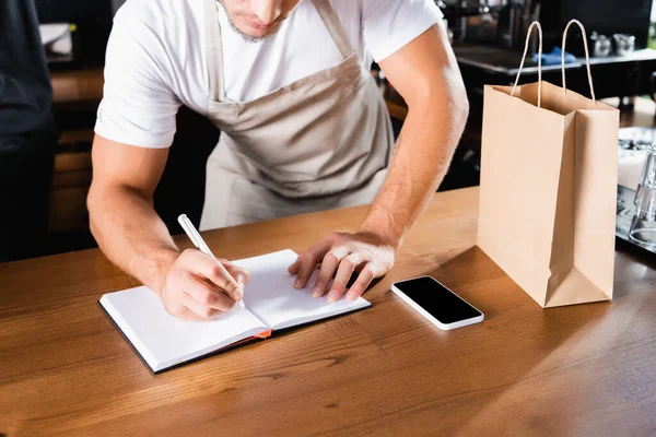 Vista ritagliata di scrittura barista in notebook vicino a sacchetto di carta e smartphone con schermo bianco sul bancone del bar — Foto stock