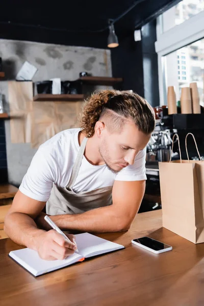 Barista looking at smartphone while writing order in notebook near paper bag — Stock Photo